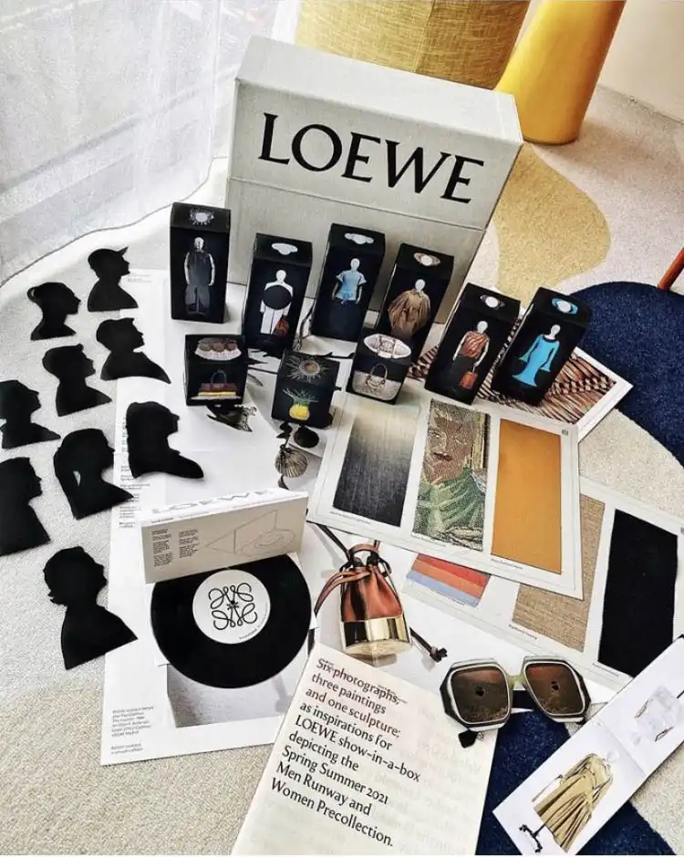 O conjunto de arquivo ‘Show in a Box’ da LOEWE disposto sobre um tapete, com as silhuetas da equipa, as caixas de roupa em miniatura, o disco de vinil e os óculos de sol